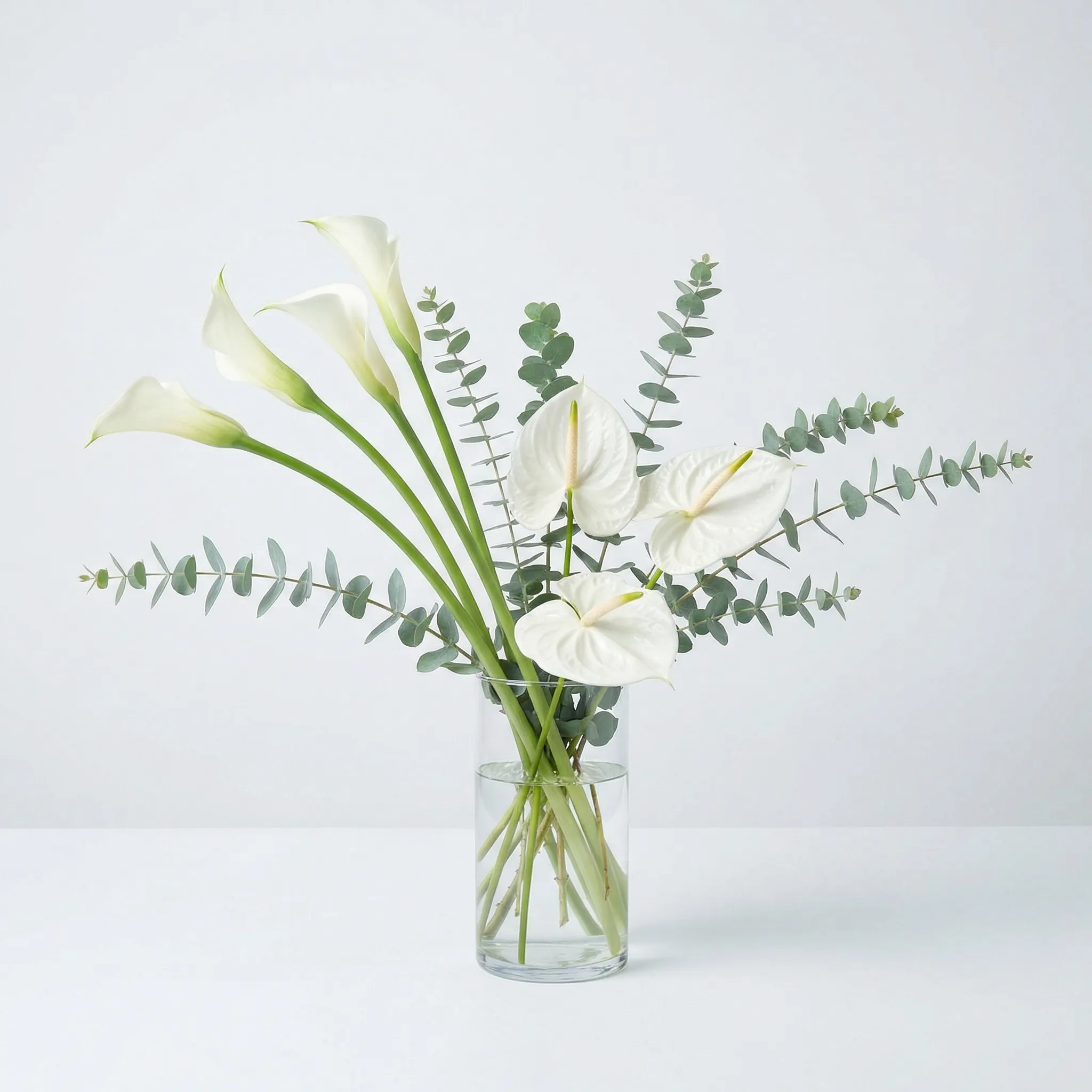 Modern minimalist arrangement with white calla lilies, anthuriums, and eucalyptus in cream and sage tones, clean studio lighting on white background