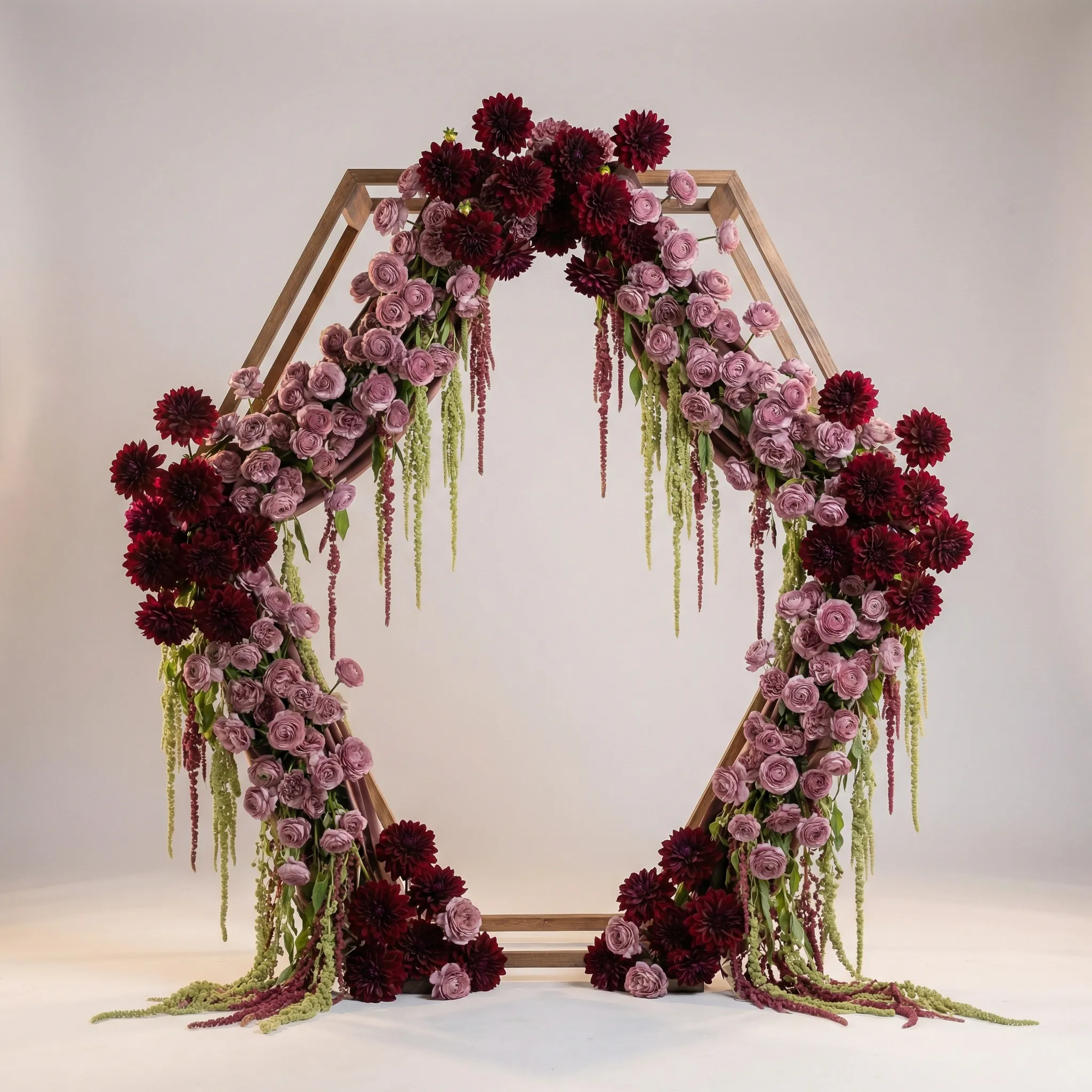 A hexagonal ceremony arch draped with burgundy dahlias, dusty mauve ranunculus, and trailing amaranthus in a romantic luxe style, soft studio lighting