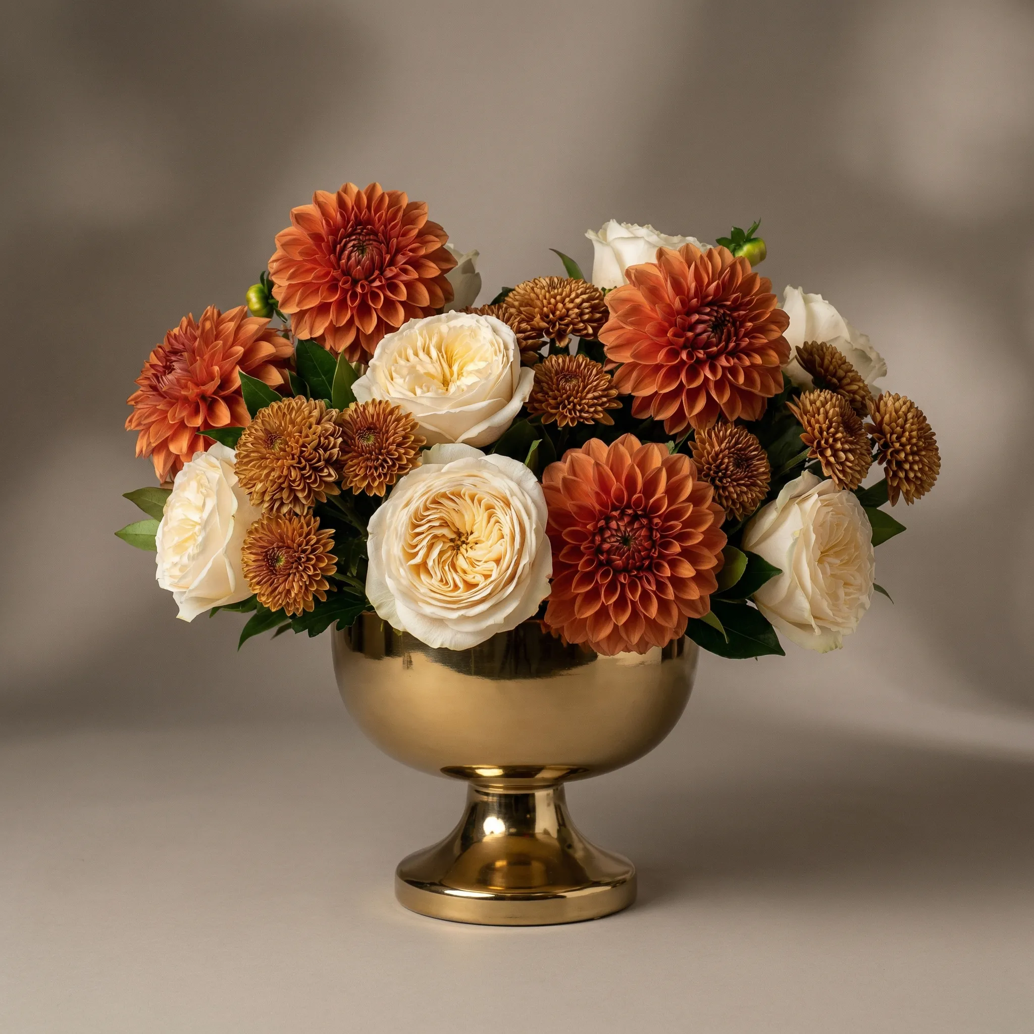 Modern centerpiece with burnt orange dahlias, cream garden roses, and bronze chrysanthemums in a gold compote vase, studio lighting