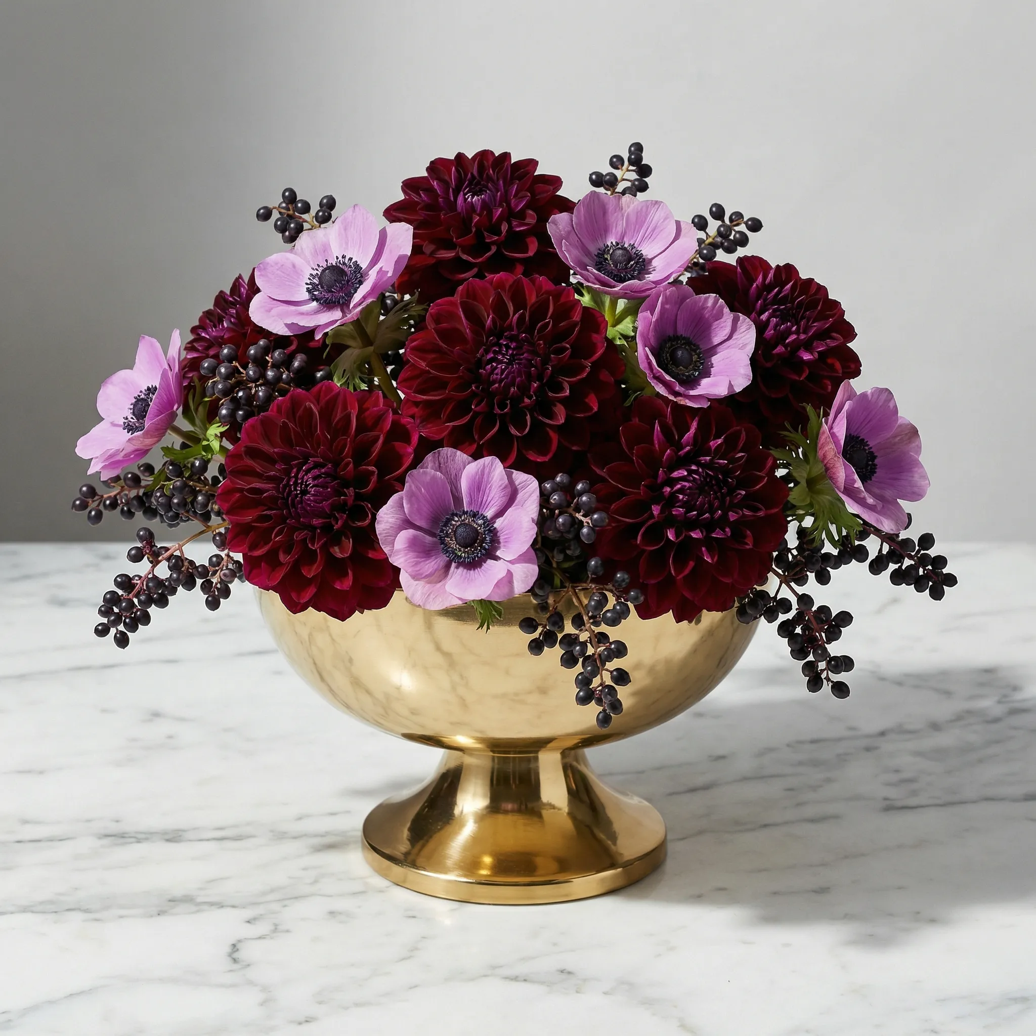 Modern geometric centerpiece with burgundy dahlias, mauve anemones, and dark privet berries in a gold compote vessel, studio lighting on marble surface